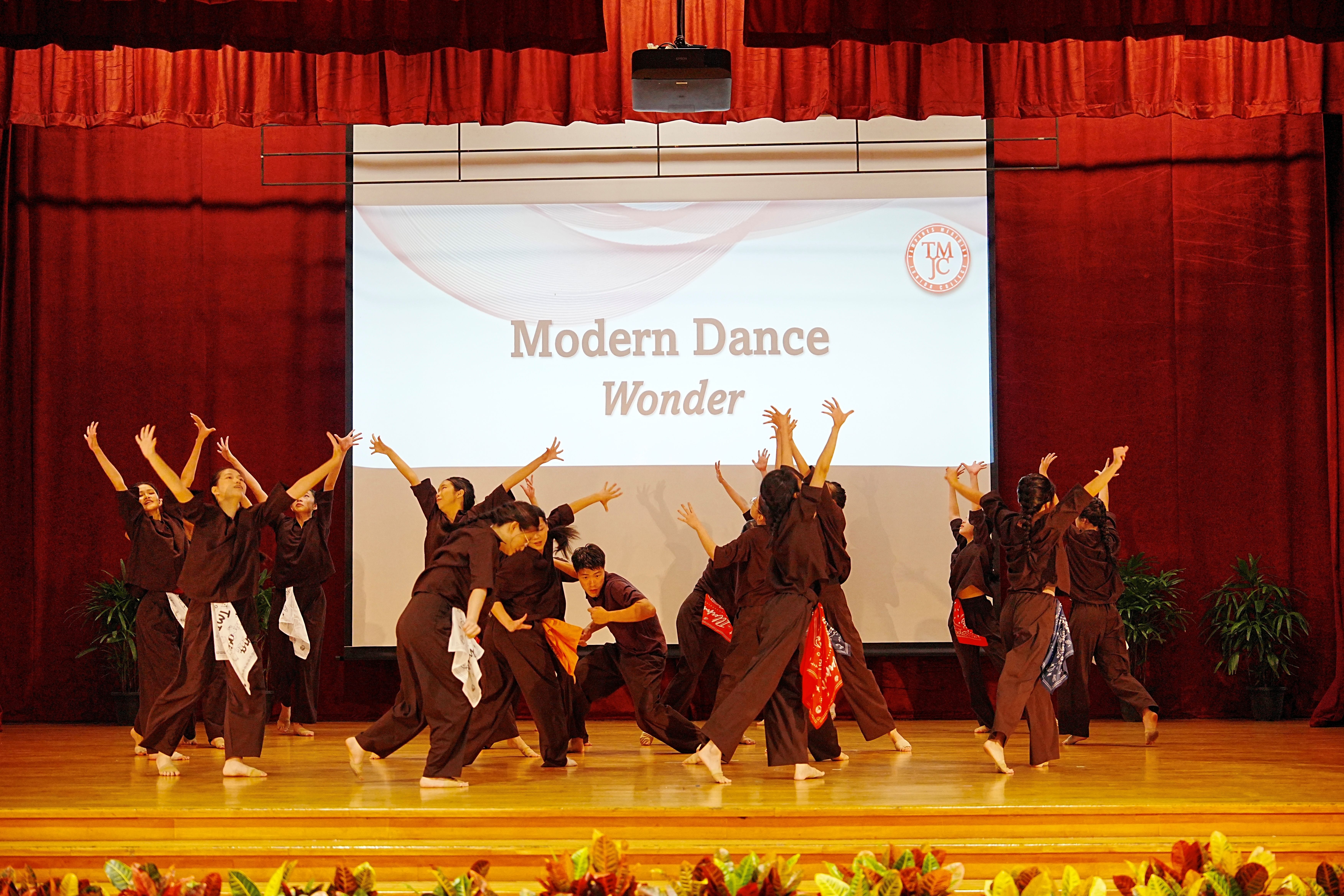 TMJC Modern Dance performers in black costumes performing 'Wonder' on stage, with arms raised expressively.